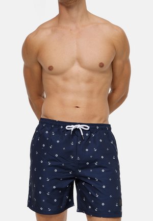 Torso masculin portant un short de bain bleu marine orné d'ancres nautiques blanches, de roues de bateau et de lettres "H", attaché avec un cordon blanc.