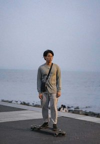 Une personne se tient sur un skateboard, portant une chemise grise à manches longues et un pantalon gris clair, avec un petit sac noir en bandoulière.
