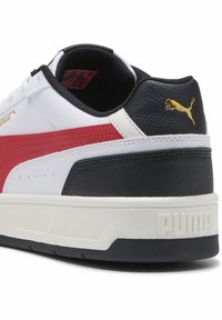 Baskets blanches avec des accents rouges et noirs, fabriquées en cuir lisse ; présentent un talon noir texturé, une semelle intermédiaire blanche et un détail de logo doré.