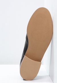 Chaussure en cuir noir avec une tige lisse et un design brogue. La semelle est en caoutchouc beige avec une surface texturée et antidérapante. Taille 37 visible.