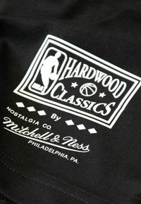 Schwarzer Stoff mit einem weißen aufgedruckten Logo, das "HARDWOOD CLASSICS" mit einer Basketballgrafik und "by Mitchell & Ness" sowie Städtenamen zeigt.