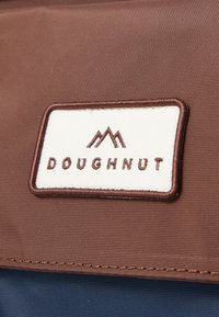 Brun etikett av stoff med "DOUGHNUT" i brun skrift på en off-white bakgrunn, med et fjelllogo over teksten. Sømmen er synlig.