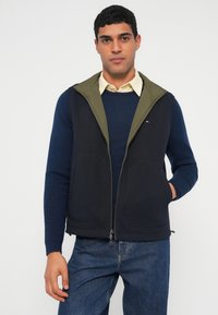 Αμάνικο γιλέκο σε ναυτικό μπλε και ελιά με φερμουάρ, δύο μπροστινές τσέπες και καπιτονέ κουκούλα, φορεμένο πάνω από μπλε πουλόβερ και πουκάμισο με γιακά.