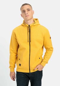 Gul zip-up hoodie med tekstureret finish, snoretræk til hætten og sorte accenter. Har to frontlommer og et logo-patch på ærmet.