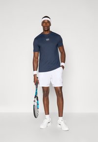 Ναυτικό αθλητικό μπλουζάκι με λογότυπο, λευκά σορτσάκια, ριγέ αθλητικοί βραχίονες και λευκά sneakers, κρατώντας ένα τένις ρακέτα με μπλε λεπτομέρειες. Απλή φόντο.