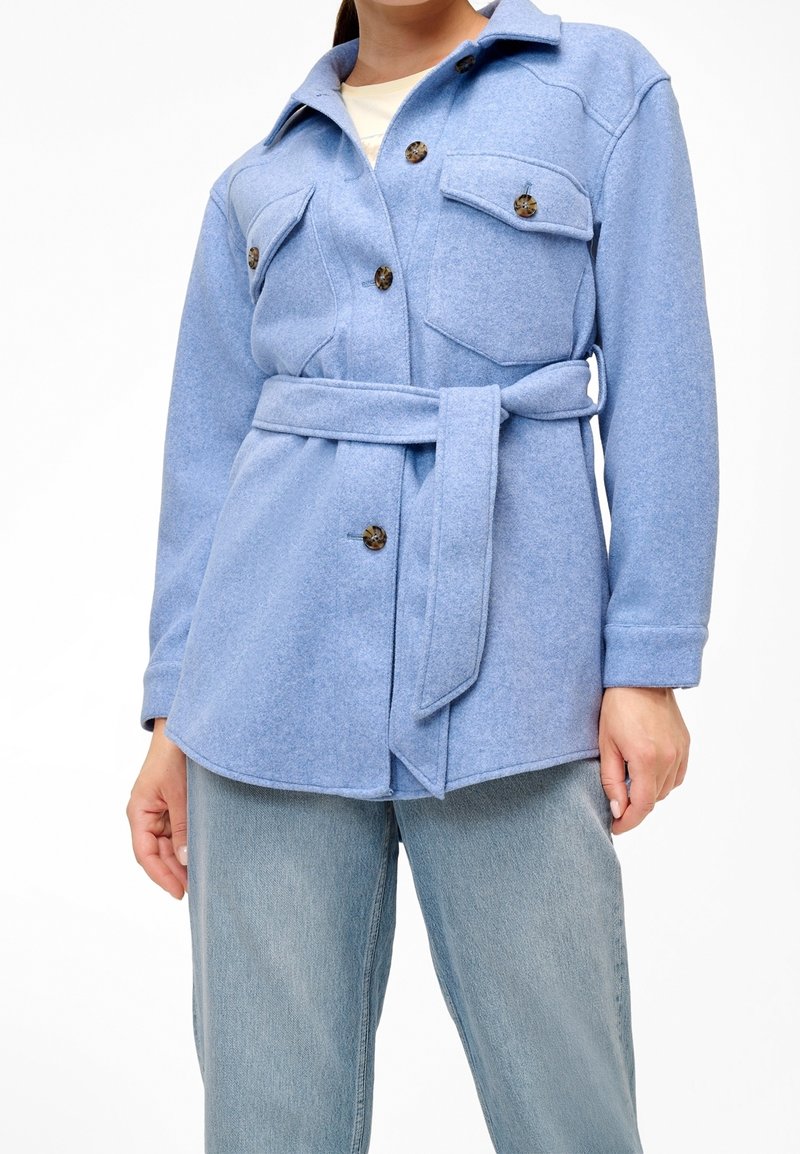 Modrá, plstěná košile-jacket s páskem v pase, s dvěma náprsními kapsami a velkými knoflíky. Nosit na světlých modrých džínách.