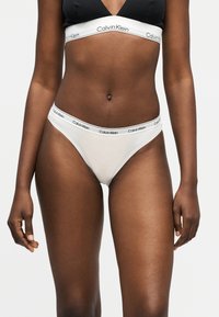 Femme portant un bralette noir Calvin Klein et un sous-vêtement blanc Calvin Klein avec des bandes élastiques de marque sur un fond uni.