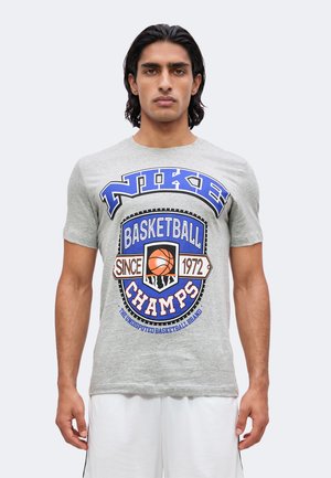 Mężczyzna w szarej koszulce Nike z grafiką o tematyce koszykówki i napisem "Basketball Champs Since 1972."