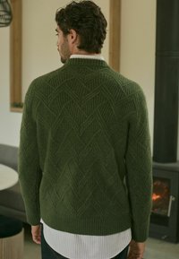 Pull en tricot vert présentant un motif de losanges texturé, des manches longues et un col montant, superposé à une chemise blanche à col.