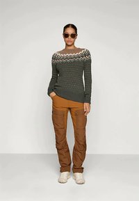Knit sweater in dark green with light brown and white geometric patterns, paired with brown cargo pants and white fuzzy slippers. Dark sunglasses worn.