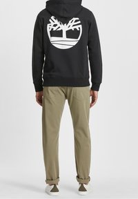 Sudadera negra con un gran logo de árbol blanco en la parte posterior, combinada con pantalones caqui y zapatos blancos. La tela parece suave y casual.