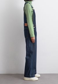 Džínové laclové kalhoty s tmavým praním, bočními kapsami, nastavovacími ramínky a béžovým koženým štítkem. Nosené přes zelený a bílý pruhovaný rolák.