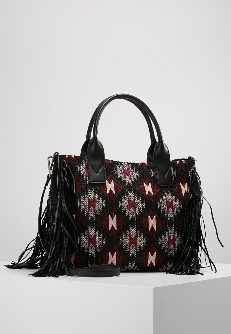 Sac à main avec des poignées en cuir noir et des franges, présentant un motif géométrique tissé en noir, bordeaux et rose sur une surface blanche.