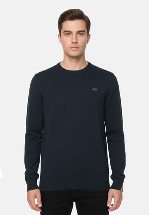 Uomo che indossa un maglione aderente a maniche lunghe blu navy con piccolo logo arancione sul petto sinistro e sul polso, su sfondo semplice.