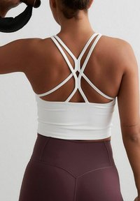 White athletic crop top featuring a strappy back design with four cross straps. Smooth texture and fitted style. Paired with dark maroon leggings.