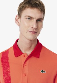 Pikétröja i orange med röd krage och en strukturerad rand. Har knappar och en grön logotyp på vänster bröst. Strukturerad tyg.