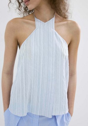 Žena nosiaca biely tielkový top bez rukávov s vertikálnymi pruhmi a modré pruhované nohavice, ruky v kapsách, smerujúca doľava.