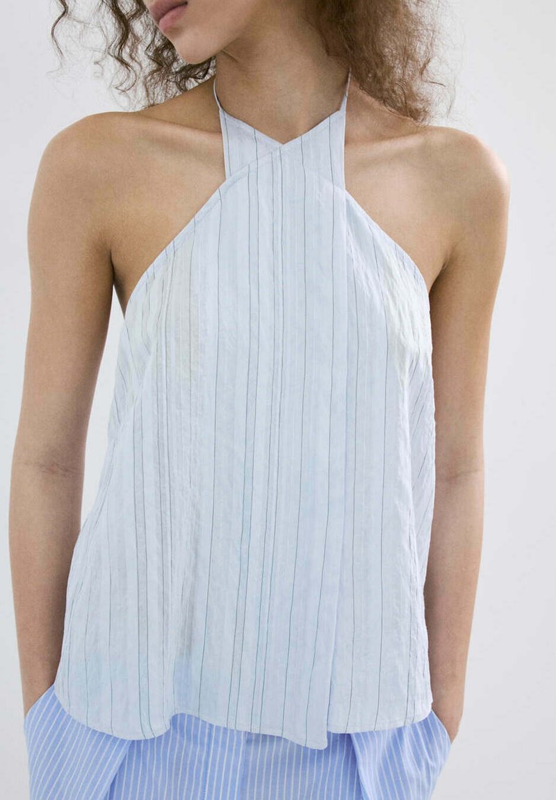 Femme portant un haut blanc sans manches à rayures verticales et un pantalon bleu rayé, mains dans les poches, regardant vers la gauche.