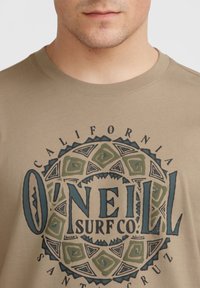 Camiseta de algodón marrón con un diseño gráfico circular en verde y azul. El texto dice "O'NEILL SURF CO." rodeado de patrones decorativos.