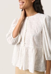 Blusa in cotone bianco con ricami floreali, scollatura rotonda, chiusura a bottoni e maniche a palloncino arricciate. L'orlo svasato aggiunge una silhouette morbida.