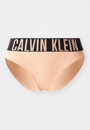 Persikų spalvos bikinio kelnaitės su juoda elastinga juosta, ant kurios didžiosiomis raidėmis parašyta "CALVIN KLEIN". Lygus tekstūros paviršius ir prigludęs dizainas.