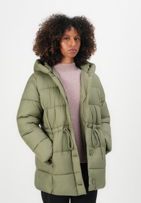 Parka verde acolchada con capucha, cintura ajustable con cordón, botones grandes y patrón de acolchado horizontal. Llevada sobre un suéter de punto acanalado rosa claro.