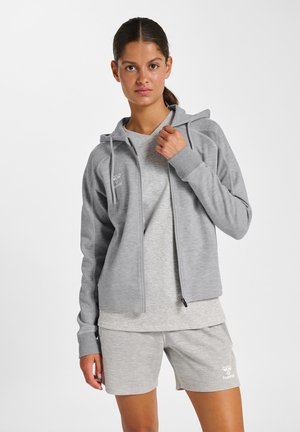 Młoda kobieta ubrana w jasnoszary kaptur, t-shirt i szorty, stoi na białym tle, trzymając zamek kaptura blisko kołnierza.