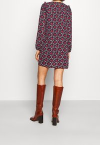 Donna che indossa un vestito a fiori corto con maniche lunghe e stivali in pelle marrone al ginocchio con tacco, in piedi davanti a uno sfondo semplice.