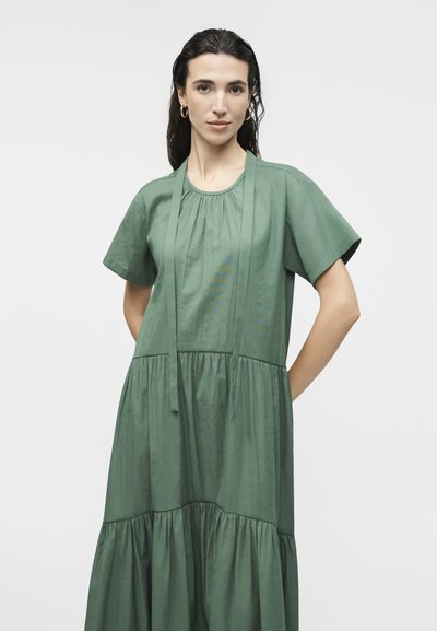 Γυναίκα με σκούρα μαλλιά φοράει φαρδιά, κοντομάνικη ρόμπα σε χρώμα φασκόμηλο με συγκεντρωμένα στρώματα και μακριές υφασμάτινες κορδέλες στον λαιμό, στέκεται μπροστά σε λευκό φόντο.