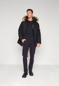 Black padded jacket with faux fur collar, paired with a black t-shirt and dark trousers. Model stands against a plain background.