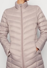 Ljusrosa puffjacka med hög krage, med chevron-uppsydda detaljer, hel dragkedja och sidofickor. Slät, vadderad textur.