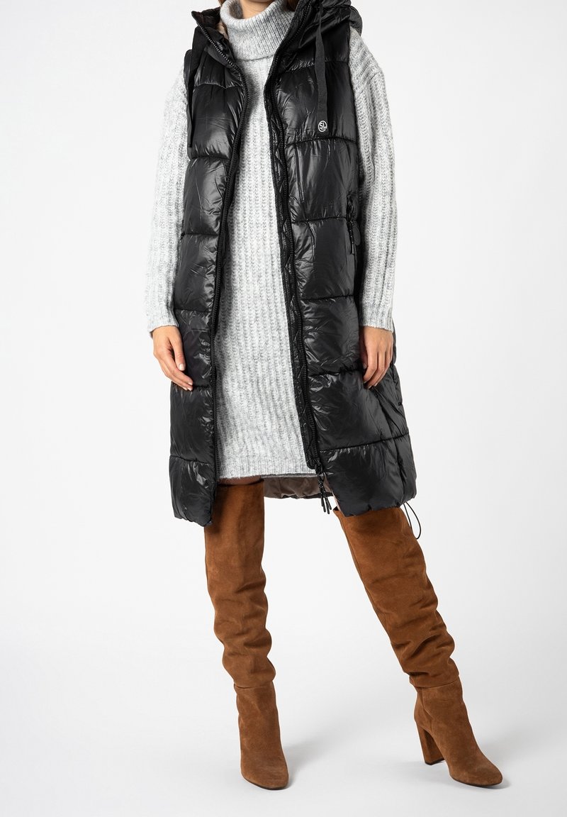 Personne portant une longue doudoune noire sans manches sur une robe en maille grise avec des bottes hautes en daim marron sur fond blanc.