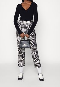 Schwarzes Langarmoberteil, hochtaillierte Hose mit Zebra-Streifen, schwarze Handtasche mit goldenen Beschlägen, schwarz-weiße Schuhe, weiße Socken.