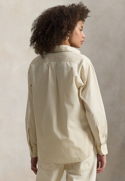 Άτομο με σγουρά μαλλιά που κοιτάζει στο πλάι, φοράει ανοιχτό μπεζ πουκάμισο με μακριά μανίκια και παντελόνι εκρού, με ουδέτερο φόντο.