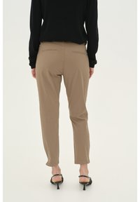 Světle hnědé kalhoty se slim fit střihem, s kapsami na zadní straně a hladkým povrchem, spárované s černými sandály na podpatku.
