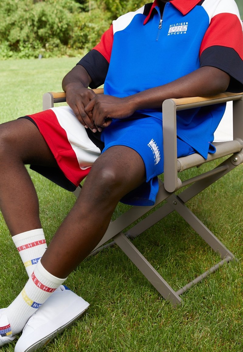 Polo à manches courtes bleu et rouge avec des accents blancs, assorti à un short bleu. Chaussettes blanches avec des bandes colorées et chaussures blanches. Assis sur une chaise sur l'herbe.