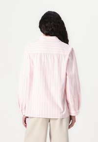 Camicia leggera a righe rosa e bianche con colletto a bottoni e maniche lunghe. Presenta una schiena plissettata e polsini con bottoni.
