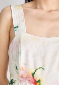 Épaule et haut de la poitrine d'une personne portant une robe sans manches avec une large bretelle plissée et un motif floral léger en aquarelle.