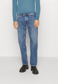 Jeans de mezclilla azul con un diseño de pierna recta, con costuras visibles, un bolsillo frontal y cierre de botón de metal. Combinados con zapatos negros.