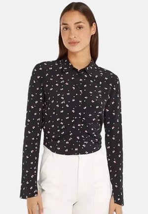 Vrouw draagt een zwart overhemd met knopen met kleine witte en roze geometrische vormen, gecombineerd met een witte broek met hoge taille.