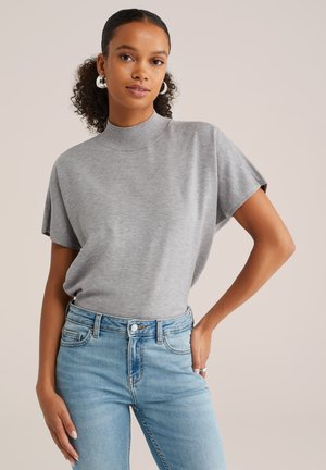 Maglietta in maglia grigia a maniche corte con scollatura alta, abbinata a jeans in denim azzurro chiaro. La maglietta ha una texture liscia ed è ampia.