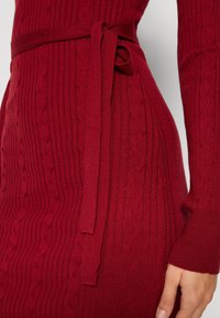 Gros plan d'une personne portant une robe en maille côtelée rouge avec une ceinture en tissu assortie nouée à la taille et des manches longues.