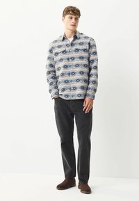 Langärmliges Hemd mit Knopfleiste aus einem gemusterten Stoff in Blau-, Beige- und Brauntönen, kombiniert mit schwarzen Hosen und braunen Stiefeln.