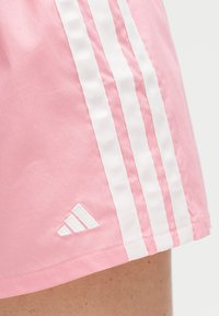 Κοντινό πλάνο ροζ αθλητικών σορτς με τρεις κάθετες λευκές ρίγες και ένα μικρό λευκό λογότυπο Adidas κοντά στο τελείωμα.