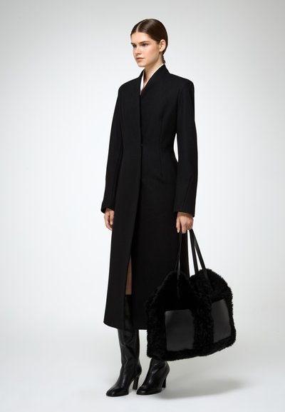 Lungo cappotto nero con colletto alto, texture liscia e spacco laterale, abbinato a stivali neri al ginocchio e una borsa nera testurizzata con dettagli in pelle.