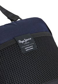 Tumši zila auduma soma ar strukturētu melnu acs paneli, uz kura ir melna birka ar brenda nosaukumu un informāciju. Regulējama siksna.