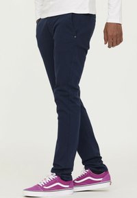 Marineblauwe slim-fit broeken met zakken en subtiele stikdetails, gecombineerd met paarse en witte low-top sneakers.