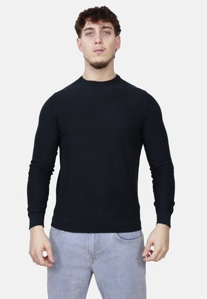Giovane uomo dalla pelle chiara e capelli ricci, indossa un maglione nero a maniche lunghe e jeans blu chiaro, su uno sfondo bianco.