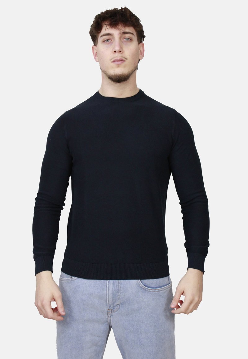 Giovane uomo dalla pelle chiara e capelli ricci, indossa un maglione nero a maniche lunghe e jeans blu chiaro, su uno sfondo bianco.