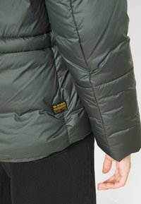 Mörkgrön puffjacka med quiltning, med en svart etikett med gul text. Tyget har en slät, glänsande struktur.